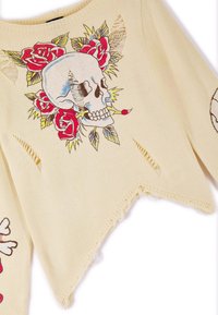Crème trui met een borduursel van een schedel en roos, met gaten voor textuur en een distressing onderrand. Korte mouwen en een losse pasvorm.