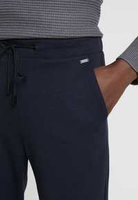 Granatowe dresy z elastycznym pasem, sznureczkiem do regulacji i boczną kieszenią. Posiadają małe srebrne logo oraz teksturowany szary materiał u góry.