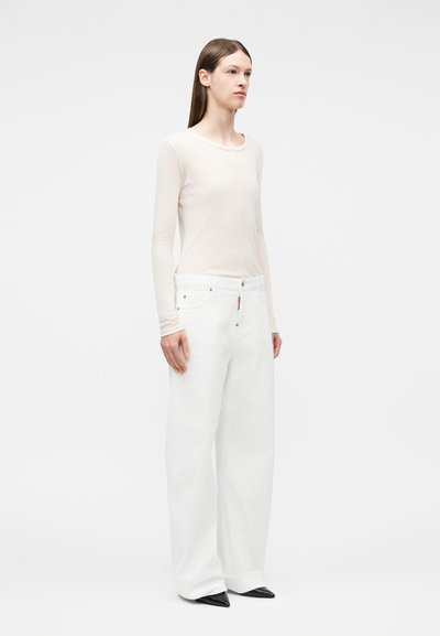 Γυναίκα που στέκεται και κοιτάζει ελαφρώς προς τα δεξιά, φορώντας εφαρμοστή μπεζ μπλούζα με μακριά μανίκια, φαρδιά λευκά παντελόνια και μαύρα μυτερά παπούτσια, σε λευκό φόντο.