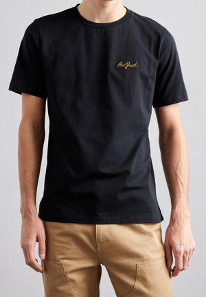 Mężczyzna w prostym czarnym t-shircie z krótkim rękawem z żółtym logo na piersi i beżowych spodniach na neutralnym tle.