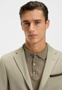 Blazer aus leicht beigefarbenem strukturiertem Stoff, einreihiges Design mit Revers, kombiniert mit einem taupefarbenen Poloshirt mit drei Knöpfen.
