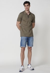 Camisa polo verde oliva con neckline de cremallera y detalles en negro, combinada con pantalones cortos de mezclilla remangados y zapatillas blancas.