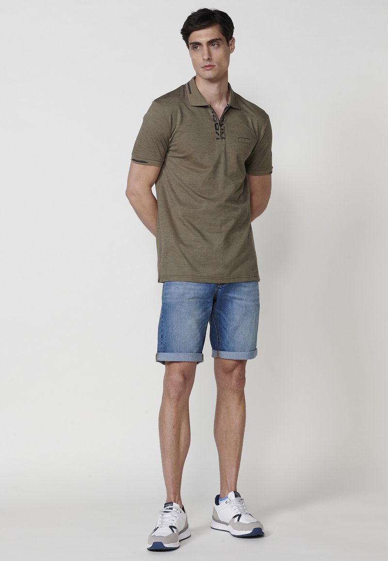 Camisa polo verde oliva con neckline de cremallera y detalles en negro, combinada con pantalones cortos de mezclilla remangados y zapatillas blancas.