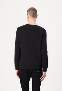 Persona con cabello corto y pendientes de aro, vistiendo una sudadera negra de manga larga y pantalones negros, de espaldas contra un fondo blanco.