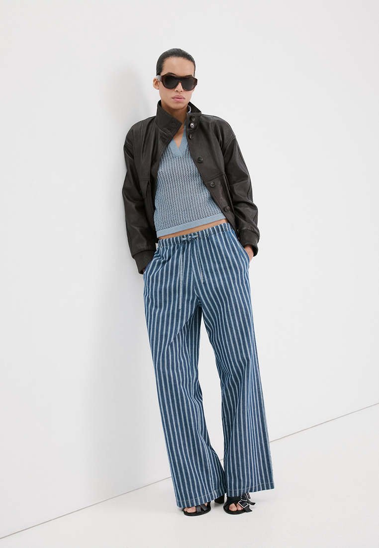 Personne portant une veste noire, un gilet en tricot bleu, un pantalon large à rayures bleues et blanches, des lunettes de soleil noires et des talons ouverts noirs, appuyée contre un mur blanc.