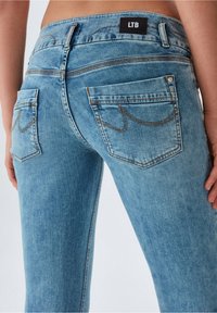 Jeans azul claro con un diseño ajustado, que cuentan con bolsillos traseros, acentos de costura marrón y una etiqueta negra "LTB" en la cinturilla.