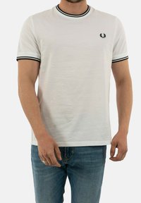 Camiseta blanca de manga corta con cuello y puños acanalados en negro, con un pequeño logo bordado en el pecho izquierdo, hecha de tela de algodón.