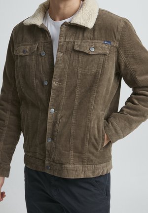 Homme portant une veste en velours côtelé marron avec un col en polaire blanche sur une chemise blanche, une main dans la poche, sur un fond uni.