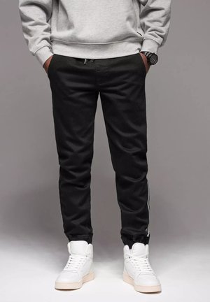 Pantalones jogger negros con corte slim, con un bolsillo con cremallera y una franja blanca en el costado. Combinados con zapatillas altas blancas.