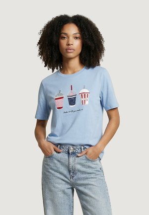 Donna con capelli ricci che indossa una maglietta azzurro chiaro con tre illustrazioni colorate di bevande e la scritta "shake it till you make it", abbinata a jeans chiari.