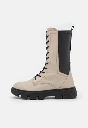 Botte de combat en cuir beige et noir avec lacets noirs, semelle crantée noire épaisse et languette à l'arrière.