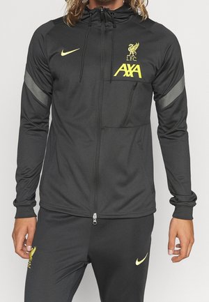 Person trägt schwarzen Liverpool FC Trainingsanzug mit gelben Nike- und AXA-Logos sowie eine Jacke mit Reißverschluss, Kapuze und reflektierenden Ärmelstreifen.