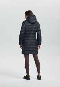 Zwarte pufferjas met een capuchon, voorzien van horizontale gestikte patronen en een middellange pasvorm. Draagt over bruine leggings en zwarte schoenen.