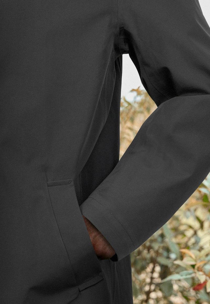 Veste imperméable noire avec une poche latérale et une texture lisse, coupe ajustée et détails de couture près des coutures.