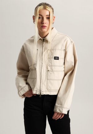 Junge Person mit langen blonden Haaren trägt eine cremefarbene Jacke mit Reißverschluss und schwarzem Vans-Logo-Patch sowie schwarze Hosen, steht vor einem einfarbigen Hintergrund.