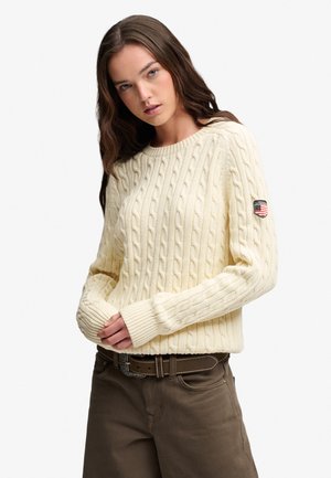 Donna che indossa un maglione crema lavorato a trecce con una toppa logo sulla manica, abbinato a pantaloni marroni e cintura, su uno sfondo neutro.