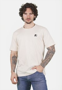 T-shirt in cotone beige con maniche corte e scollatura rotonda. Presenta un piccolo logo nero "Starter" sul lato sinistro del petto. Vestibilità casual.