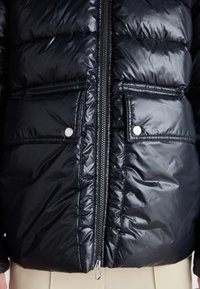 Casaco puffer em material preto brilhante, apresentando um fecho de correr na frente, dois bolsos exteriores com botões de pressão e uma textura acolchoada.