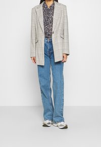 Overdimensioneret ternet blazer i beige og sort, parret med en blomstermønstret navy skjorte, højtaljede blå jeans og hvide sneakers.