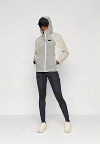 Lichtgrijze en beige jas met capuchon en ritssluiting, gecombineerd met marineblauwe leggings. Voorzien van een gestructureerd ontwerp en een getextureerde stof.