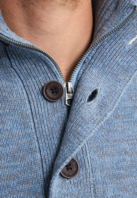 Primer plano de un suéter de punto azul con la cremallera parcialmente subida y botones marrones, visible en el pecho y el cuello de una persona.