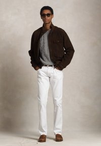 Chaqueta de ante marrón, suéter gris de punto trenzado, jeans blancos y zapatos marrones. Modelo con gafas de sol oscuras de pie con las manos en los bolsillos.