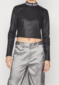 Czarna, skórzana bluzka z długim rękawem i krótkim krojem z wysokim kołnierzem, z białym logo "Calvin Klein". Połączona z błyszczącymi srebrnymi spodniami.