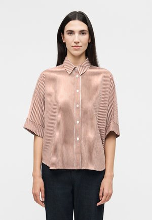 Mujer con cabello lacio y oscuro lleva una camisa de manga corta, suelta, abotonada, con rayas verticales rojas y blancas y pantalones oscuros.