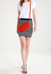 Borsa a tracolla rossa con una texture liscia e forma curvata, abbinata a una t-shirt bianca e una gonna a righe blu e bianche. Sandali argentati.