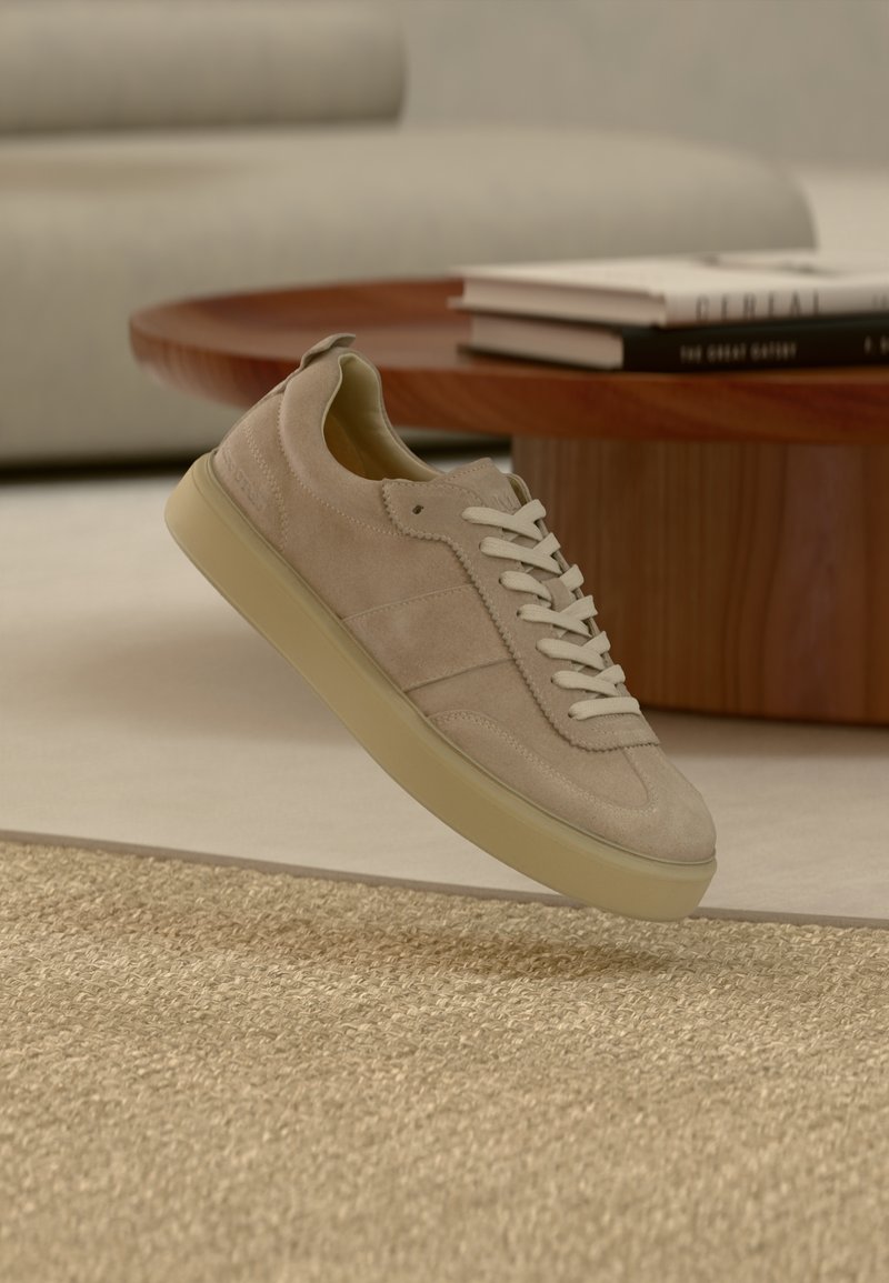 Μπεζ σουέντ αθλητικό παπούτσι με λευκά κορδόνια να επιπλέει πάνω από μπεζ χαλί με υφή, ξύλινο στρογγυλό τραπέζι και βιβλία σε ουδέτερο σαλόνι.