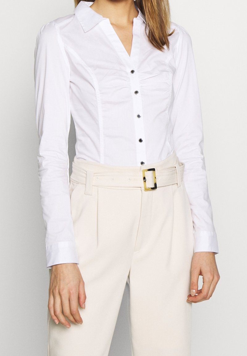 Vit åtsittande skjorta med krage, långa ärmar och svarta knappar, tillsammans med krämfärgade högmidjade byxor och ett bälte med guldspänne.