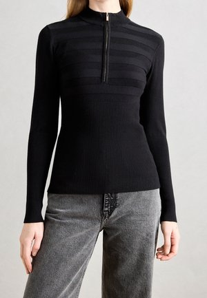 Frau trägt einen körpernahen schwarzen, gerippten Pullover mit horizontalen Streifen und einem Frontreißverschluss, kombiniert mit grauen Jeans aus Denim.