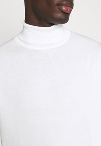Maglione bianco a collo alto realizzato in un tessuto morbido, caratterizzato da un design aderente e un collo a coste. Texture liscia senza motivi visibili.