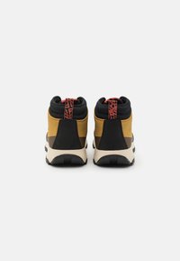 Las zapatillas de caña alta presentan una combinación de materiales negros, amarillos y marrones con un acabado texturizado y un acento decorativo rojo en la parte trasera.