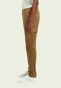 Ruskotaiset corduroy cargo-housut, joissa on sivutaskut, slim fit -leikkaus ja teksturoitu kangas, yhdistettynä valkoisiin tennareihin. Sivunäkymä.