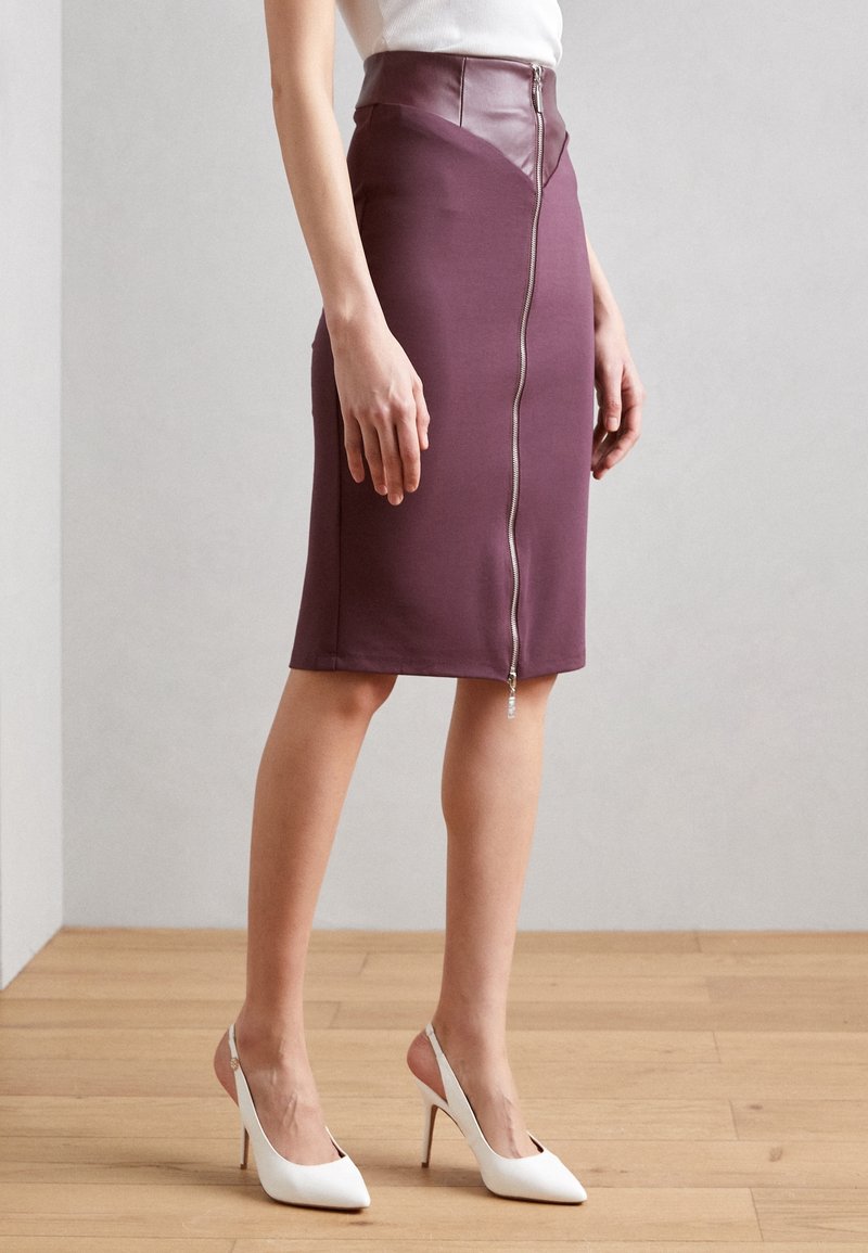 Figurbetonter, knielanger Rock aus tiefviolettem Stoff mit einem ledernen Bund und einem Reißverschlussdetail auf der Vorderseite. Getragen mit weißen High-Heels.