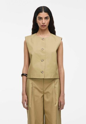 Kvinde med langt mørkt hår, iført ærmeløs beige knap-op-top og matchende brede bukser, stående mod en enkel hvid baggrund.