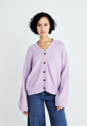 Lichtpaarse gebreide cardigan met een V-hals, zes houten knopen en ribgebreide mouwen, gecombineerd met blauwe wijde jeans.