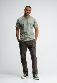 Man in een olijfgroene t-shirt, donkere broek en zwarte sneakers staat met zijn handen in zijn zakken tegen een witte achtergrond.