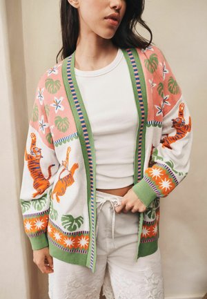 Frau trägt eine bunte Strickjacke mit Tiger-, Blumen- und Blattmustern über einem weißen Crop-Top und weißen Spitzen-Shorts.