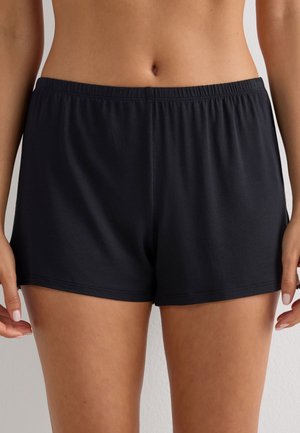 Kvinde iført løstsiddende sorte elastikshorts, der er beskåret midt på låret, stående mod en ensfarvet lys baggrund.