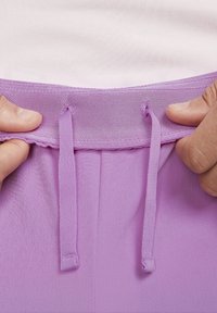 Händer som håller och drar åt lavendelfärgade snörning på ett lila midjeband över en ljusrosa texturerad tyg.