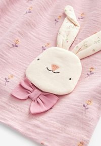 Tissu rose à motifs floraux orné d'une application tête de lapin avec un nœud plié en rose doux, créant un design texturé et ludique.