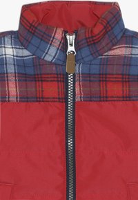 Czerwona ocieplana kurtka z niebieskim i czerwonym nadrukiem w kratkę na kołnierzu i górnej części klatki piersiowej, wyposażona w centralny srebrny zamek błyskawiczny z brązowym uchwytem.