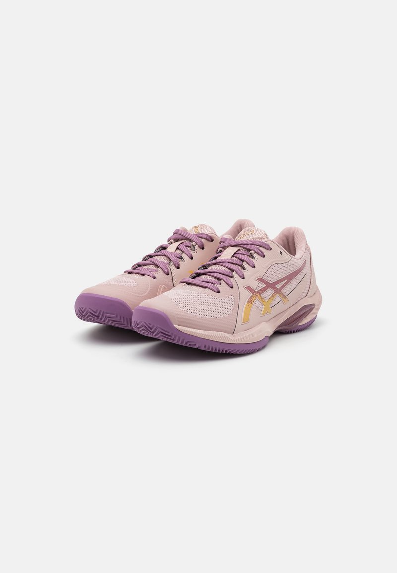 Zapatillas deportivas de color rosa claro con acentos púrpuras, parte superior de malla, suela de goma gruesa y detalle del logo dorado. Diseño de punta redondeada con cierre de cordones.