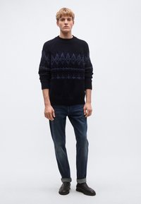 Tengerészkék kötött pulóver geometriai mintákkal a mellrészen, sötét farmernadrággal és fekete csúszásmentes cipővel párosítva. A modell világos háttér előtt áll.