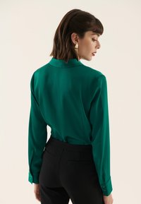 Blusa verde a maniche lunghe in tessuto liscio, con colletto e polsini con bottoni, abbinata a pantaloni aderenti neri.