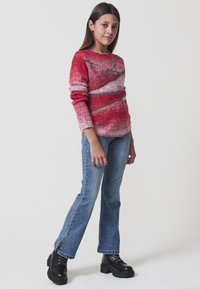 Maglione a righe rosse e rosa con maniche lunghe, abbinato a jeans a zampa blu e stivaletti neri con lacci.