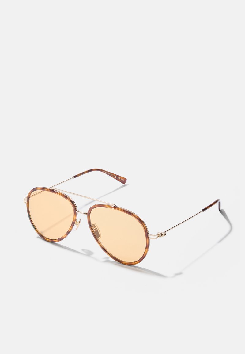Gafas de sol marrones, en forma ovalada, con lentes de tono ámbar, montura de metal ligera y un sutil patrón en las varillas. Brazos delgados y curvados.