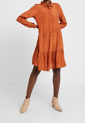 Frau trägt ein locker sitzendes, orangefarbenes, langärmliges Kleid mit Knöpfen und beigefarbene Stiefeletten, steht mit einer Hand in der Hüfte.